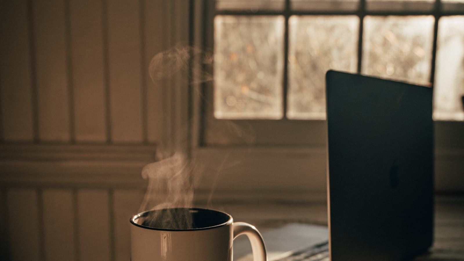 Steaming mug of tea beside an open laptop by a window